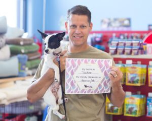 A man holds a small black and white dog in one arm and a sign in the other that reads, My name is Tucker. My family thinks Im a terrier/chi. They are inside a pet store with shelves of pet supplies behind them.