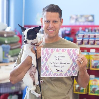 A man holds a small black and white dog in one arm and a sign in the other that reads, My name is Tucker. My family thinks Im a terrier/chi. They are inside a pet store with shelves of pet supplies behind them.